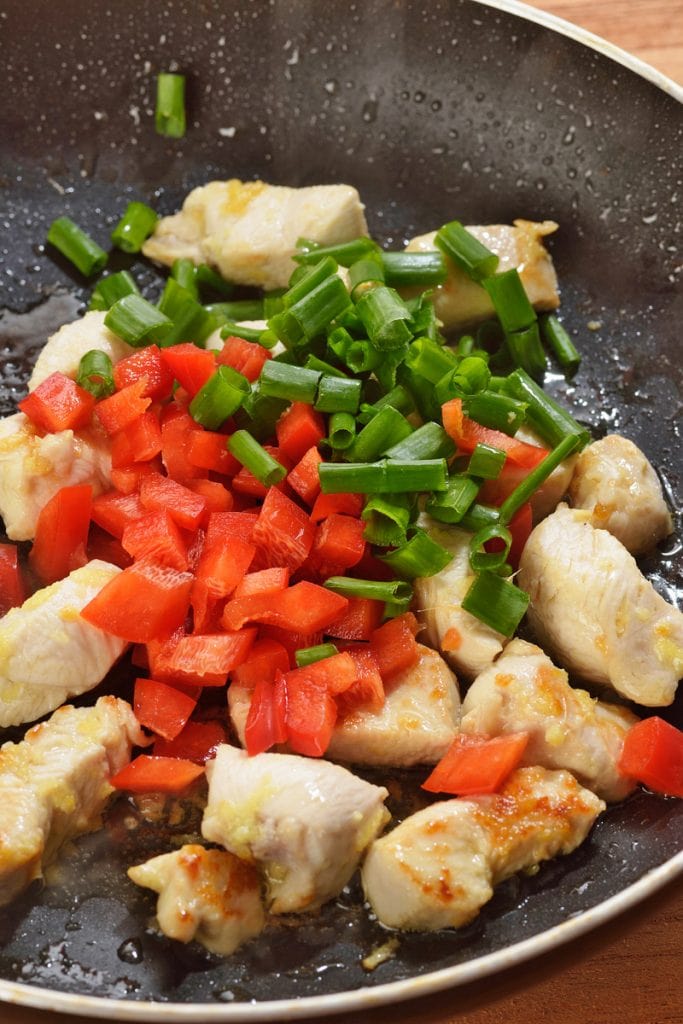 Adding red bell peppers, spring onion, and chicken to a pan or wok
